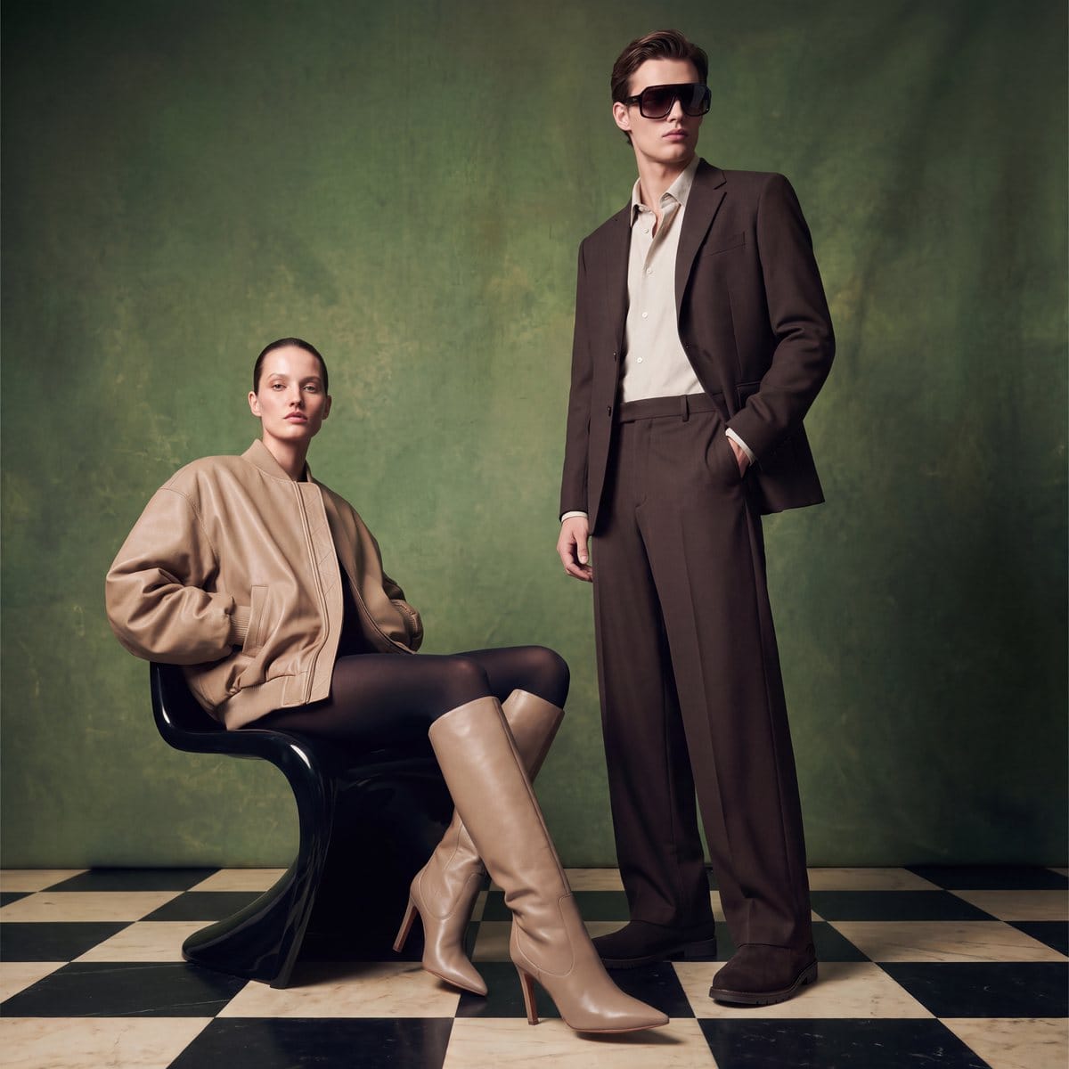 Two models in brown tones against a green backdrop and checkerboard floor