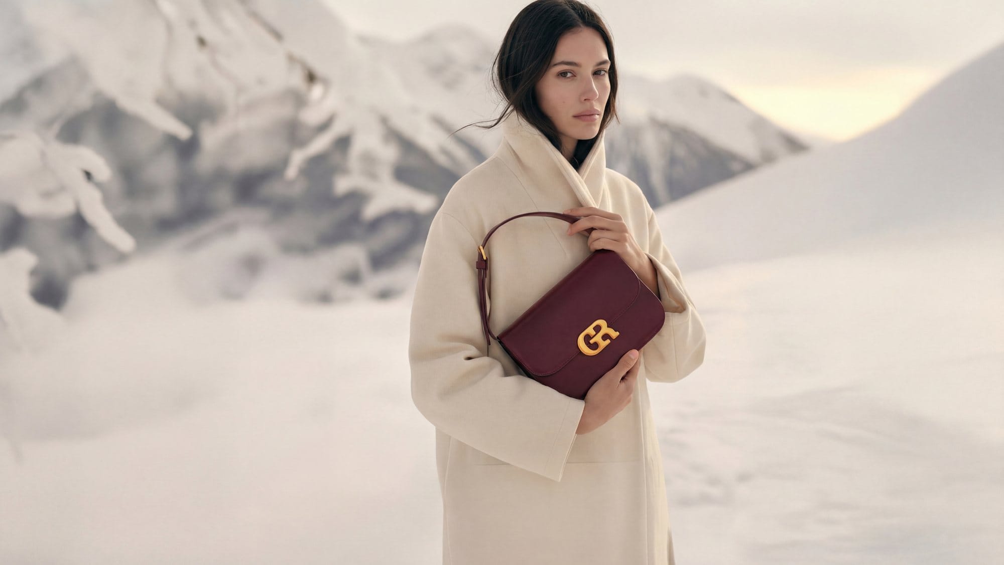 Model in a cream coat with a burgundy bag against snowy mountains